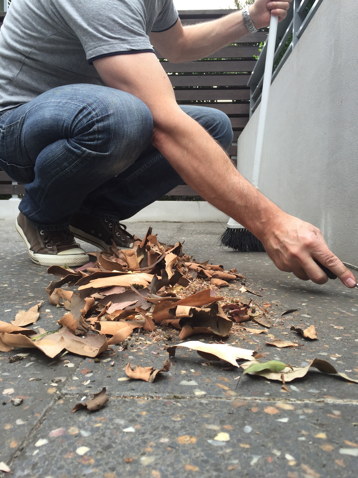Husband in jeans, t-shirt and sneakers crouching cleaning sweeping leaves terrazzo tiled backyard