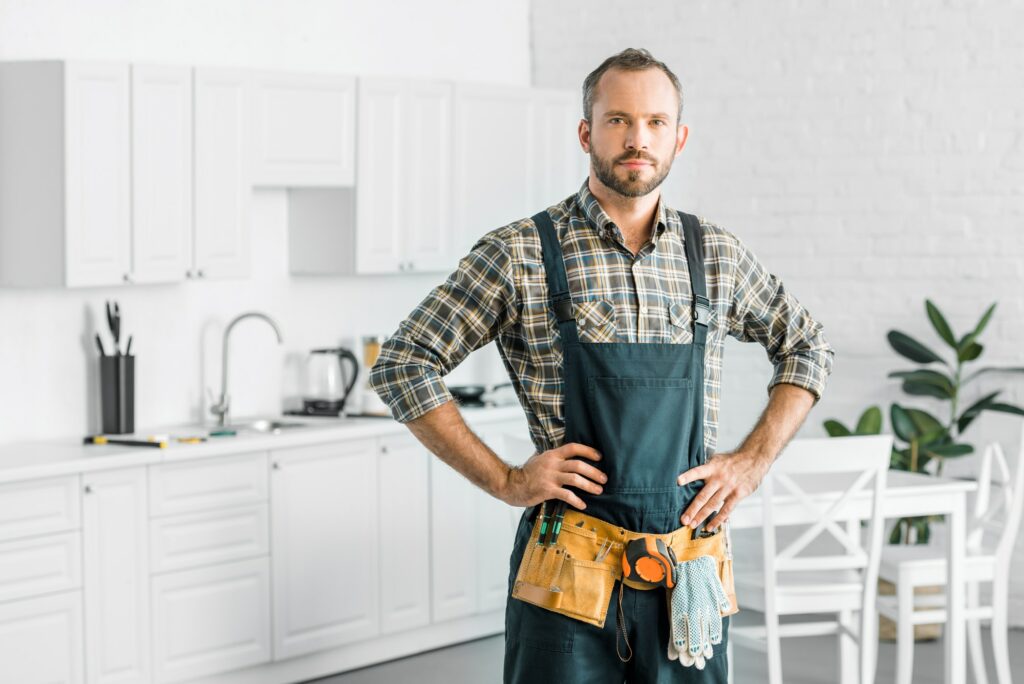 handsome plumber in overalls and tool belt looking at camera in kitchen